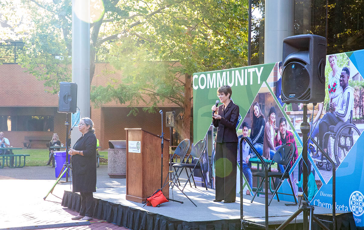 President Howard on stage in the new quad of the Salem Campus during Fall Kickoff events. She is holding a microphone and talking. A podium is in front of her, behind her is a banner with images of students and the word 